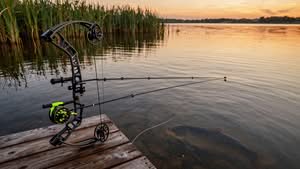 A compound bow with a fishing reel is resting on a dock by a lake at sunset, with a large fish visible in the water near the dock.