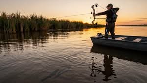 A person stands on a boat in shallow water at sunset, aiming a bow and arrow at fish visible beneath the water's surface near tall reeds.