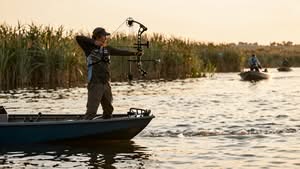 A person stands on a boat in a marsh, aiming a compound bow at the water; other people in boats are visible in the background.